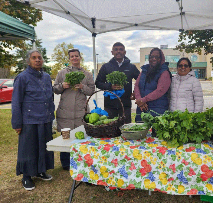A photo of Community & Family Program Services Manager Amy Semenuk. She is wearing a tan winter coat and holding a bouquet of basil. She is joined by several other volunteers in similar winter attire, standing in front of a table of bitter melon, celery, okra and misc. herbs and greens.