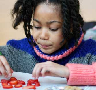 A young girl is sitting at a table eating a spread of sliced strawberries in front of her. She’s wearing a blue, pink, and grey turtleneck sweater with her hair in two pig's tails.