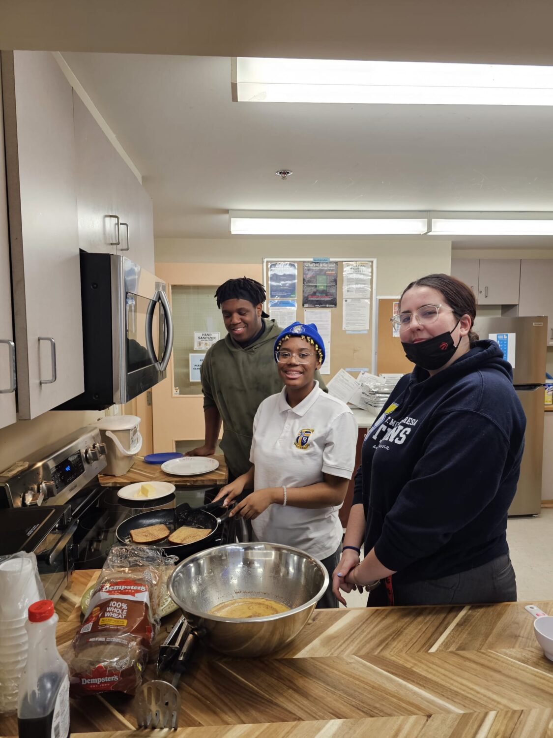 three participants, one male presenting and two females presenting smile in front of the stove as they prepare French toast for the youth cooking program. Two of these individuals are members of the Malvern Lead Program.