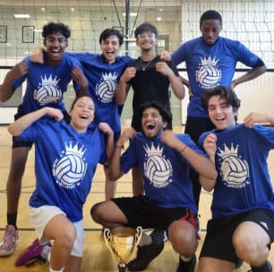 seven male presenting youth participants in matching blue shirts with a crown and volleyball emblem, posing in front of gym volleyball net at 90 Littles Rd.