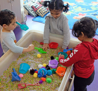 Three children in the EarlyOn program are playing in a sandbox together with shovels and toys. Two girls one boy all of them are standing. The boy has short brown hair and is wearing a grey long sleeve shirt with black pants. Both girls have two pig tails in their hair. Girl on the right is wearing red sweater with black pants and the girl in the middle is wearing a grey sweater.