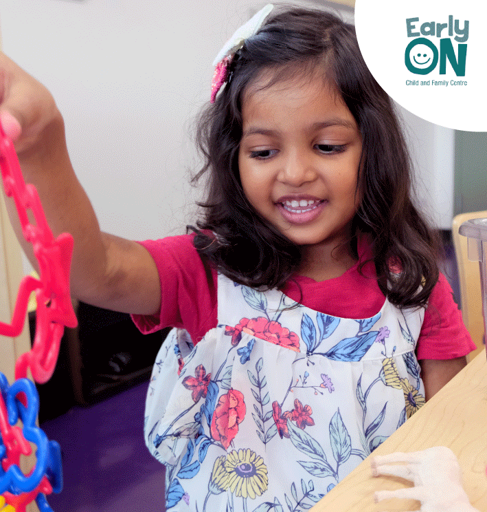 South Asian Girl presenting smiling while playing with a toy , wearing a white and red floral dress.