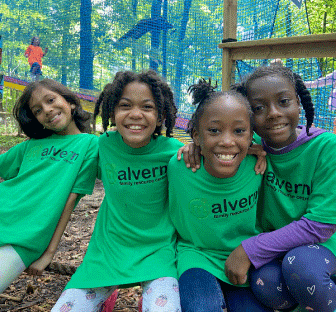 There are four young girls wearing bright green Malvern Family Resource centre t-shirts. They are sitting closely together, engaged in a lively conversation smiling and sharing joyful moments with each other outside.