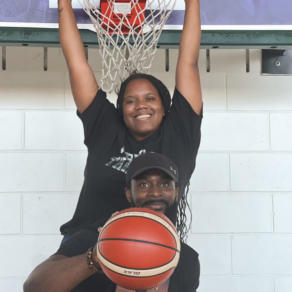 Shaniqua Wright, youth development worker stands near the basketball rim, holding onto it, dressed in a black logo tee. Recreational Program Coordinator Randy Blackwood poses in front of her, with a basketball in hand, dressed in a black graphic tee and a black hat.