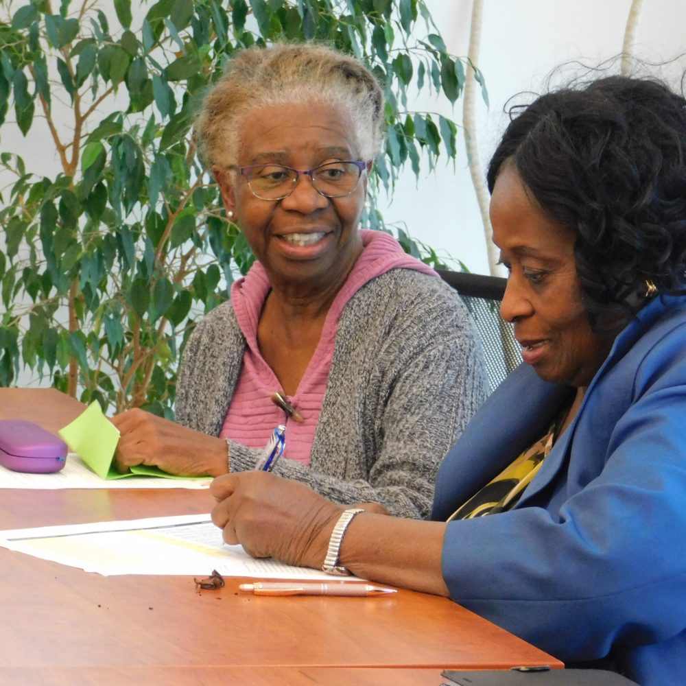 Two senior women of African American* descent. It’s a candid photo as neither woman seems to be aware of the camera. The one on the left has glasses and is wearing a pink sweater with a grey cardigan over it. The woman to the right is wearing a blue blazer and has a silver watch on her left wrist.